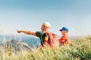 Père et fils assis dans l’herbe au sommet, admirant le lac du Bourget et les montagnes de Savoie.